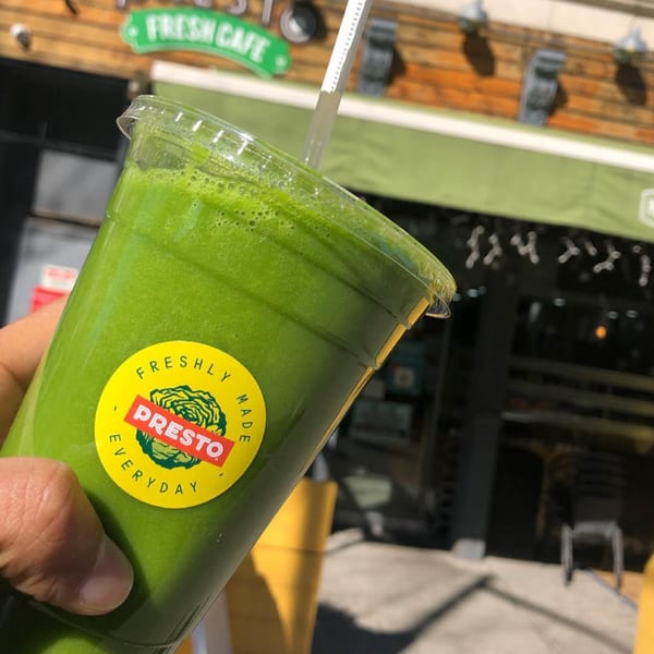 Hand holding a green smoothie with a "Presto Freshly Made Everyday" sticker, outside the cafe.